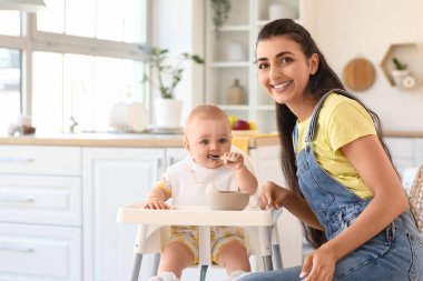 Genç anne bebeğini evdeki yüksek sandalyede besliyor.