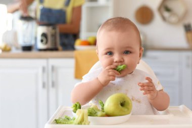 Yüksek sandalyede brokoli yiyen şirin bir bebek. Annesinin evde püre hazırlamasına karşı. Yakın plan.