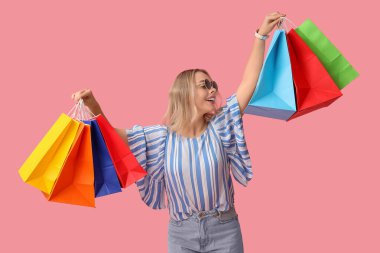 Pembe arka planda alışveriş torbaları olan güneş gözlüklü genç bir kadın.