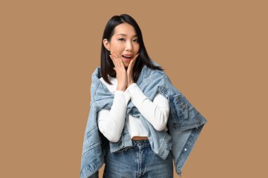 Kahverengi arka planda şık kot elbiseli güzel, genç, şok olmuş Asyalı bir kadın.