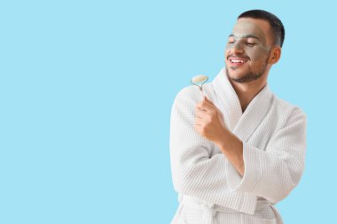 Yüzünde kozmetik jel maskesi ve mavi arka planda masaj maskesiyle bornozlu yakışıklı genç adam.