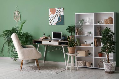 Diz üstü bilgisayarı, raf ünitesi ve evdeki yeşil duvarın yanındaki farklı ev bitkileriyle şık bir iş yeri.