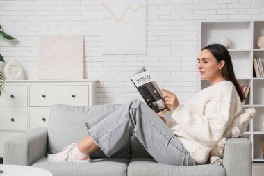 Evdeki kanepede iç tasarım dergisi okuyan genç bir kadın.