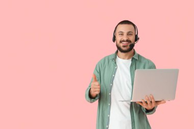 Kulaklıklı, dizüstü bilgisayarlı genç adam pembe arka planda baş parmağını gösteriyor.