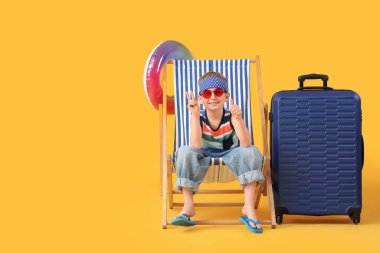 Sevimli küçük çocuk, sarı arka planda şezlongda oturan şişirilebilir yüzük ve bavulla zafer jesti gösteriyor. Seyahat kavramı