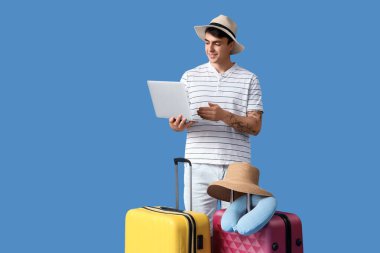 Mavi arka planda dizüstü bilgisayar kullanan bavullu bir erkek turist.