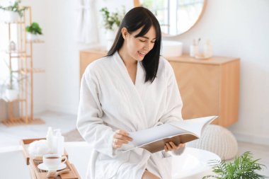 Evde bornoz giymiş, dergi okuyan genç bir kadın.