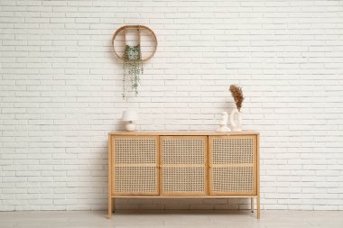 Modern hafif ahşap çekmeceler içinde lamba ve vazoda beyaz tuğla duvarın yanında otlar var.