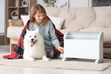 Ekoseli ve Samoylu köpekli genç bir kadın radyatörün yanında ısınıyor.
