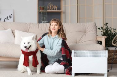 Ekoseli ve Samoylu köpekli genç bir kadın radyatörün yanında ısınıyor.