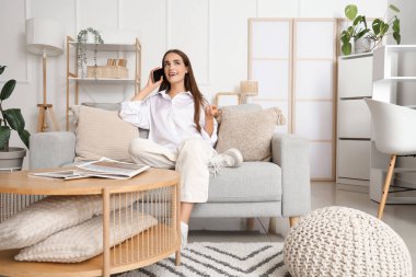 Aydınlık oturma odasındaki gri koltukta cep telefonuyla konuşan genç bir kadın.