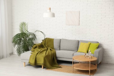 Asılı lamba, ekoseli yumuşak kanepe, çiçek, resim ve sehpa beyaz tuğla duvarın yanında.