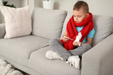 Evdeki kanepede oturan termometresi ve mendili olan hasta çocuk.