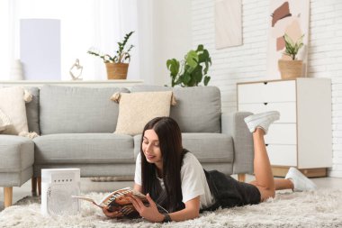 Portatif vantilatörü olan, yerde uzanan ve oturma odasında dergi okuyan güzel genç bir kadın.