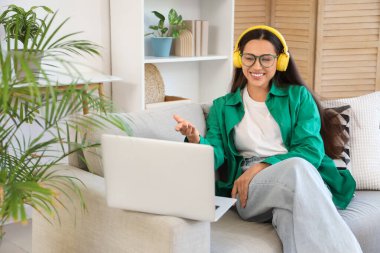 Kulaklıklı, dizüstü bilgisayarlı, evdeki kanepede sohbet eden güzel bir genç kadın.