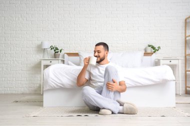 Yakışıklı genç adam yerde oturuyor ve yatak odasında çay içiyor.