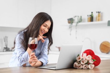 Şarap içen ve mutfakta erkek arkadaşıyla görüntülü sohbet eden genç bir kadın. Online randevu