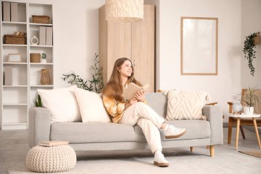 Güzel genç bir kadın şık oturma odasındaki kanepede kitap okuyor.