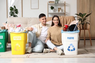 Çöpleri çöp tenekelerine dizilmiş, evde yerde oturan genç bir çift. Küresel Geri Dönüşüm Günü