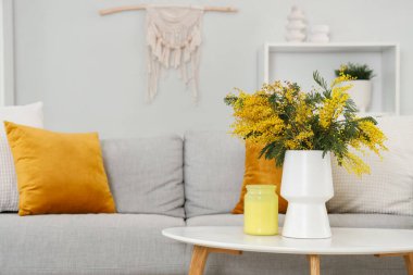 Oturma odasındaki kanepenin yanındaki masada mimoza çiçekleri olan vazo.