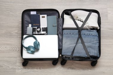 Gri ahşap zeminde giysi ve modern aletlerle dolu bavulu aç.