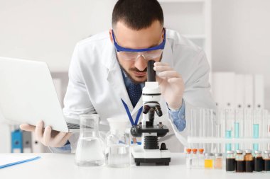 Laboratuvarda mikroskopla inceleyen dizüstü bilgisayarı olan erkek bilim adamı.