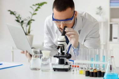 Laboratuvarda mikroskopla inceleyen dizüstü bilgisayarı olan erkek bilim adamı.