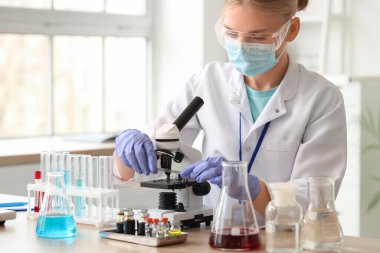 Tıbbi maskesi ve mikroskobu olan kadın bilim adamı laboratuarda çalışıyor.