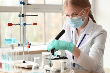 Tıbbi maskesi ve mikroskobu olan kadın bilim adamı laboratuarda çalışıyor.