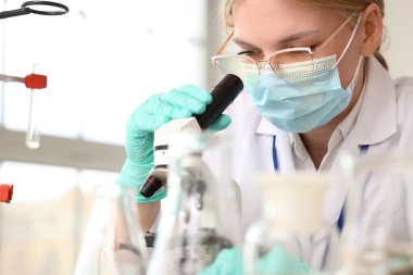 Tıbbi maskeli bir kadın bilim adamı mikroskopla laboratuara bakıyor.