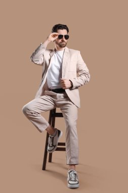 Açık renk takım elbiseli, güneş gözlüklü, bej arka planda, taburede oturan yakışıklı bir genç.