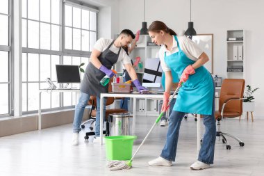 Kadın hademe yerleri paspaslıyor ve ekibi ofisi temizliyor.