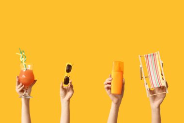 Sarı arka planda güneş kremi, kokteyl ve güneş gözlüklü mini iskemle tutan kadın eller.