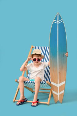 Sörf tahtası olan şirin bir çocuk mavi arka planda iskemlede oturuyor. Seyahat kavramı