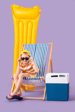 Güverte sandalyesinde oturan şirin küçük çocuk şişme şiltesi ve leylak arka planda plaj soğutucusuyla. Seyahat kavramı