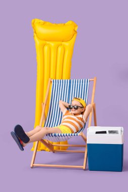 Güverte sandalyesinde oturan şirin küçük çocuk şişme şiltesi ve leylak arka planda plaj soğutucusuyla. Seyahat kavramı