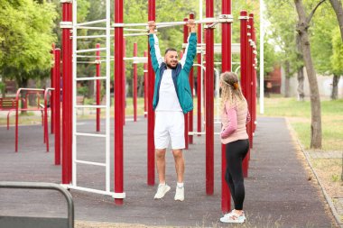 Antrenör, genç bayana parkta nasıl barfiks çekileceğini gösteriyor.
