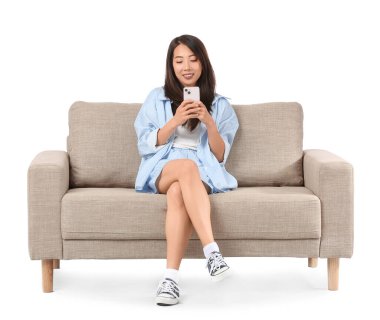 Genç Asyalı kadın gri koltukta beyaz arka plana karşı cep telefonu kullanıyor.