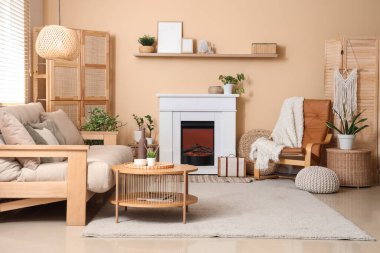 Oturma odasının iç tarafındaki bej duvarın yanında şömine, kanepe, masa ve deri koltuk.