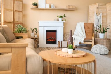 Oturma odasının iç tarafındaki bej duvarın yanında şömine, kanepe, masa ve deri koltuk.