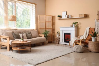 Oturma odasının iç tarafındaki bej duvarın yanında şömine, kanepe, masa ve deri koltuk.