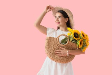 Yazlık şapkalı şık genç kadın elinde hasır çantasıyla pembe arka planda ayçiçekleri tutuyor.