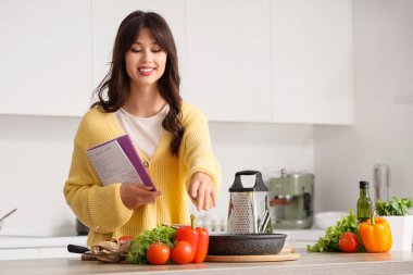 Yemek tarifi kitabıyla mutfakta yemek pişiren genç bir kadın.