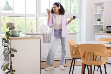 Kulaklıklı, kahve ve cep telefonlu, mutfakta şarkı söyleyen Afro-Amerikalı genç bir kadın.