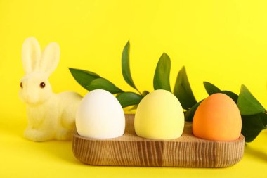 Boyanmış Paskalya yumurtaları, yeşil dalları ve sarı arka planda tavşanı olan tahta tutucu.