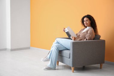 Genç Afro-Amerikalı kadın turuncu duvarın yanındaki gri koltukta dergi okuyor.
