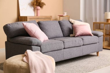 Oturma odasındaki bej duvarın yanında yastıklı, puflu ve şifoniyerli kanepe.