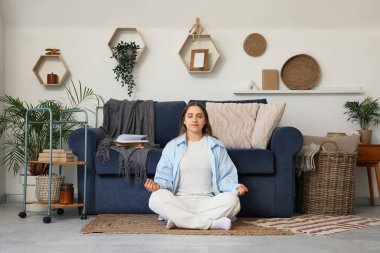 Genç bir kadın oturma odasındaki mavi kanepenin yanında meditasyon yapıyor.