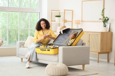 Cep telefonlu genç Afro-Amerikalı kadın evdeki kanepede seyahat için bavul hazırlıyor.