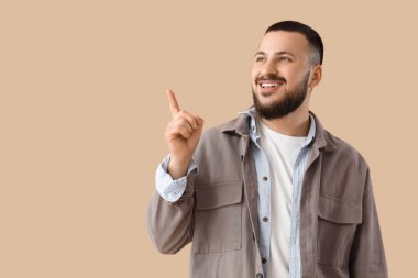 Güzel gülüşü olan genç adam bej arka plandaki bir şeyi işaret ediyor.